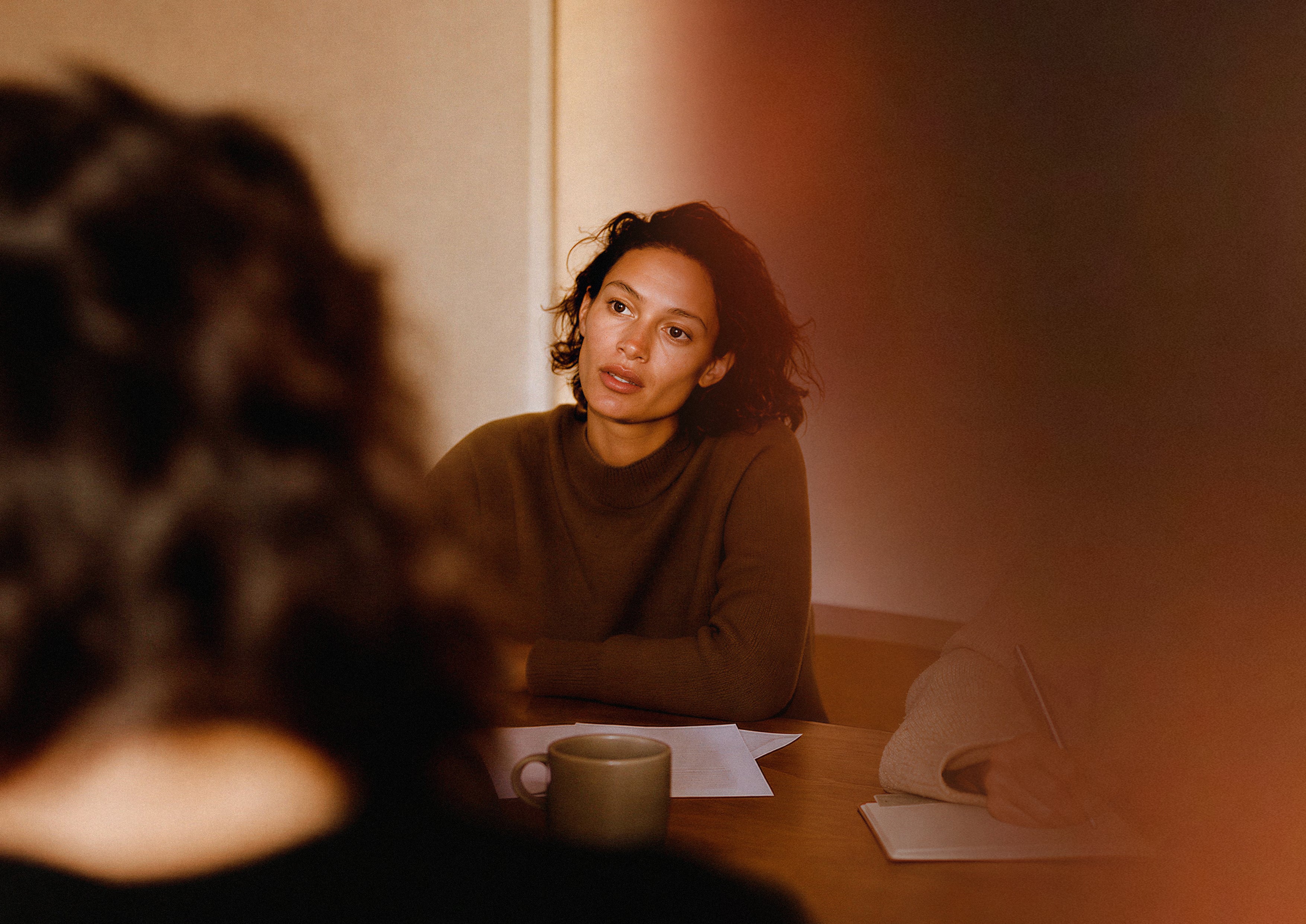 A woman with dark hair sits at a wooden table, looking thoughtfully to the side in a warmly lit indoor setting. Papers and a mug are on the table, with a blurred figure in the foreground.