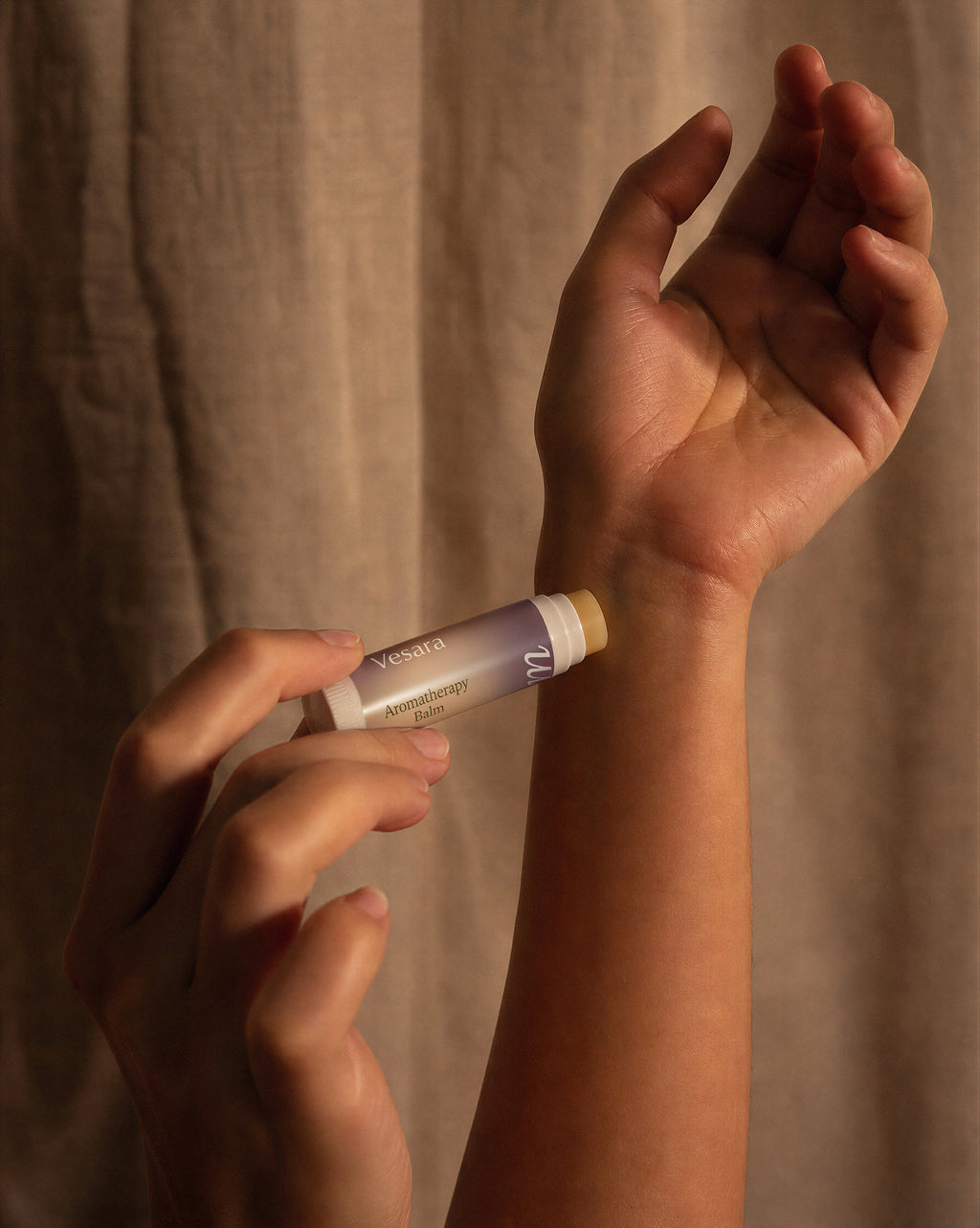 A close-up of a person applying a tube of Vesara Calm aromatherapy balm to their wrist, with a beige fabric curtain in the background.
