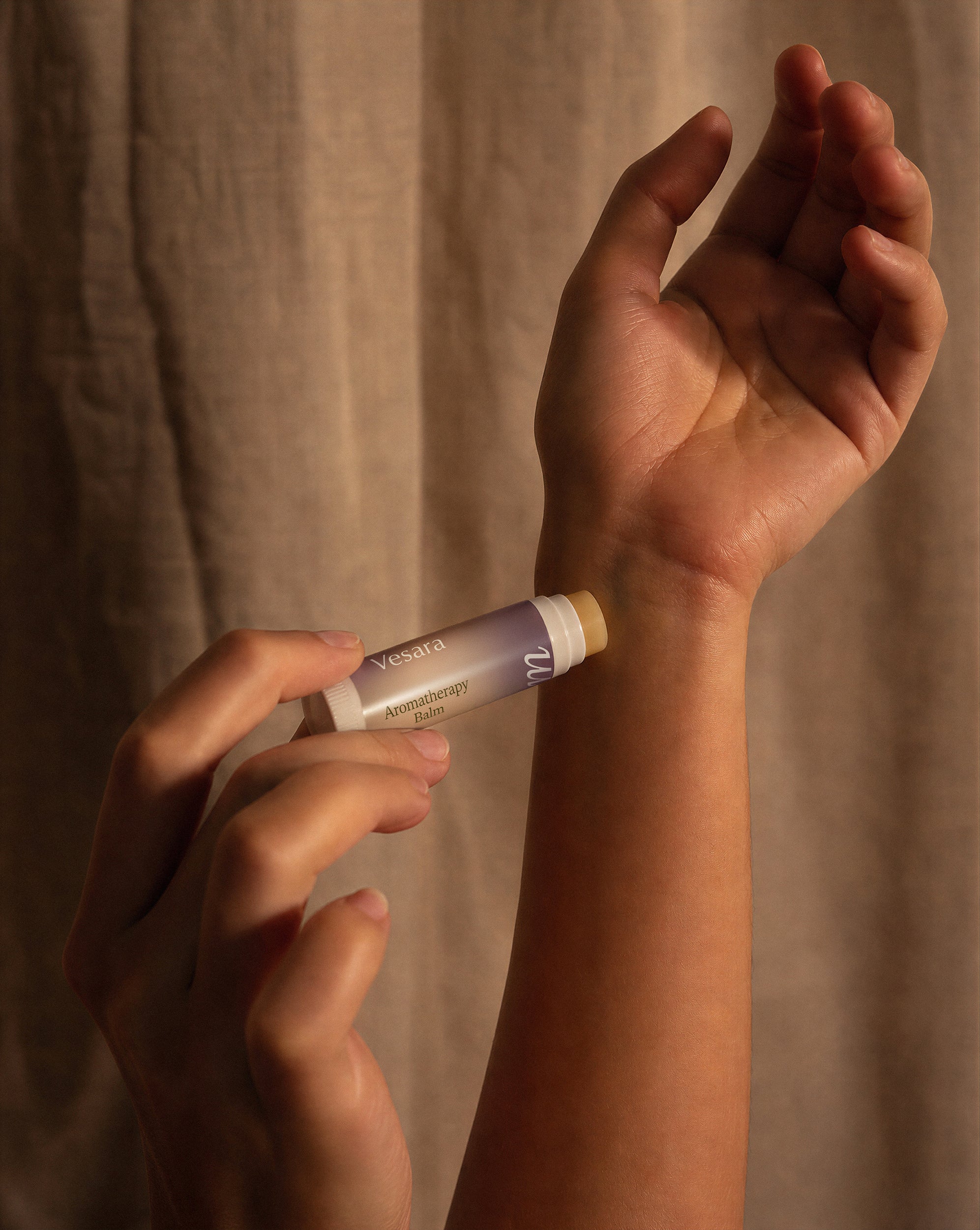 A close-up of a person applying a tube of Vesara Calm aromatherapy balm to their wrist, with a beige fabric curtain in the background.