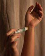A close-up of a person applying a tube of Vesara Clarity aromatherapy balm to their wrist, with a beige fabric curtain in the background.
