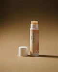 Vesara Energize Aromatherapy Balm tube standing upright with white cap removed and sitting off to the left, both standing on warm beige surface