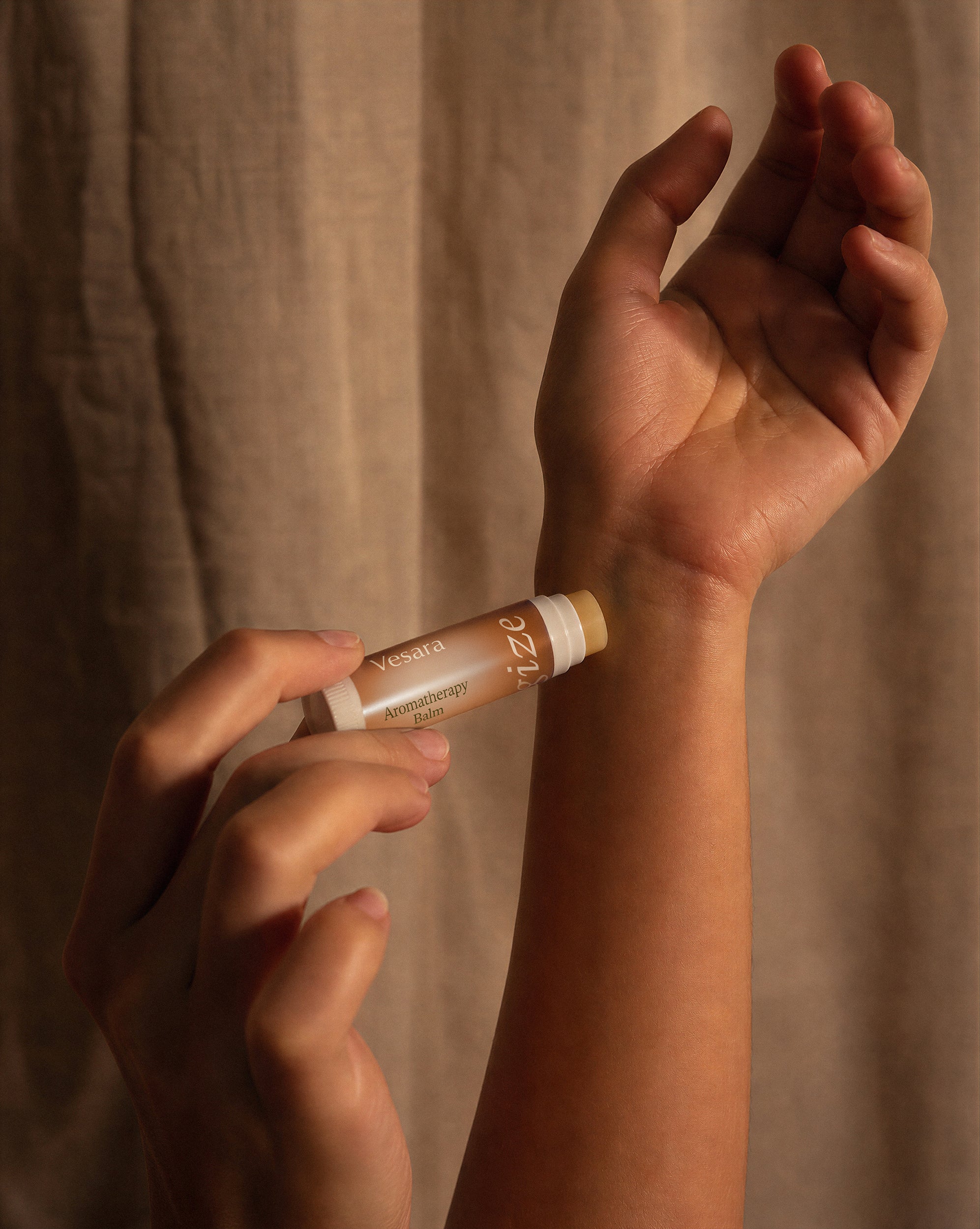 A close-up of a person applying a tube of Vesara Energize aromatherapy balm to their wrist, with a beige fabric curtain in the background.