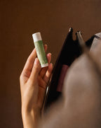 A hand holding Vesara aromatherapy balm clarity tube with white cap next to an open purse with a warm brown background