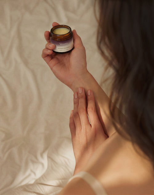 Looking over a woman's shoulder, she has a jar of Vesara's Calm Aromatic Hand Salve in one hand with the cap off revealing the salve inside, the other hand is rubbing the product into her wrist, soft studio lighting