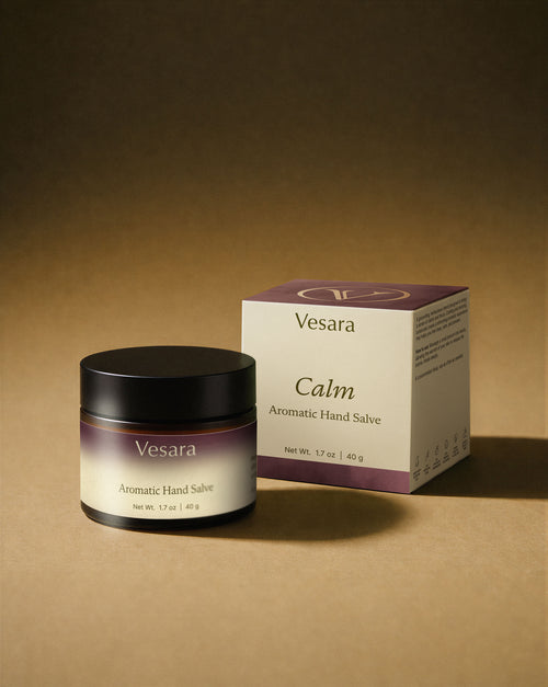 Vesara Calm hand salve round amber glass jar with a black lid and packaging standing together against a warm beige background with soft moody studio lighting