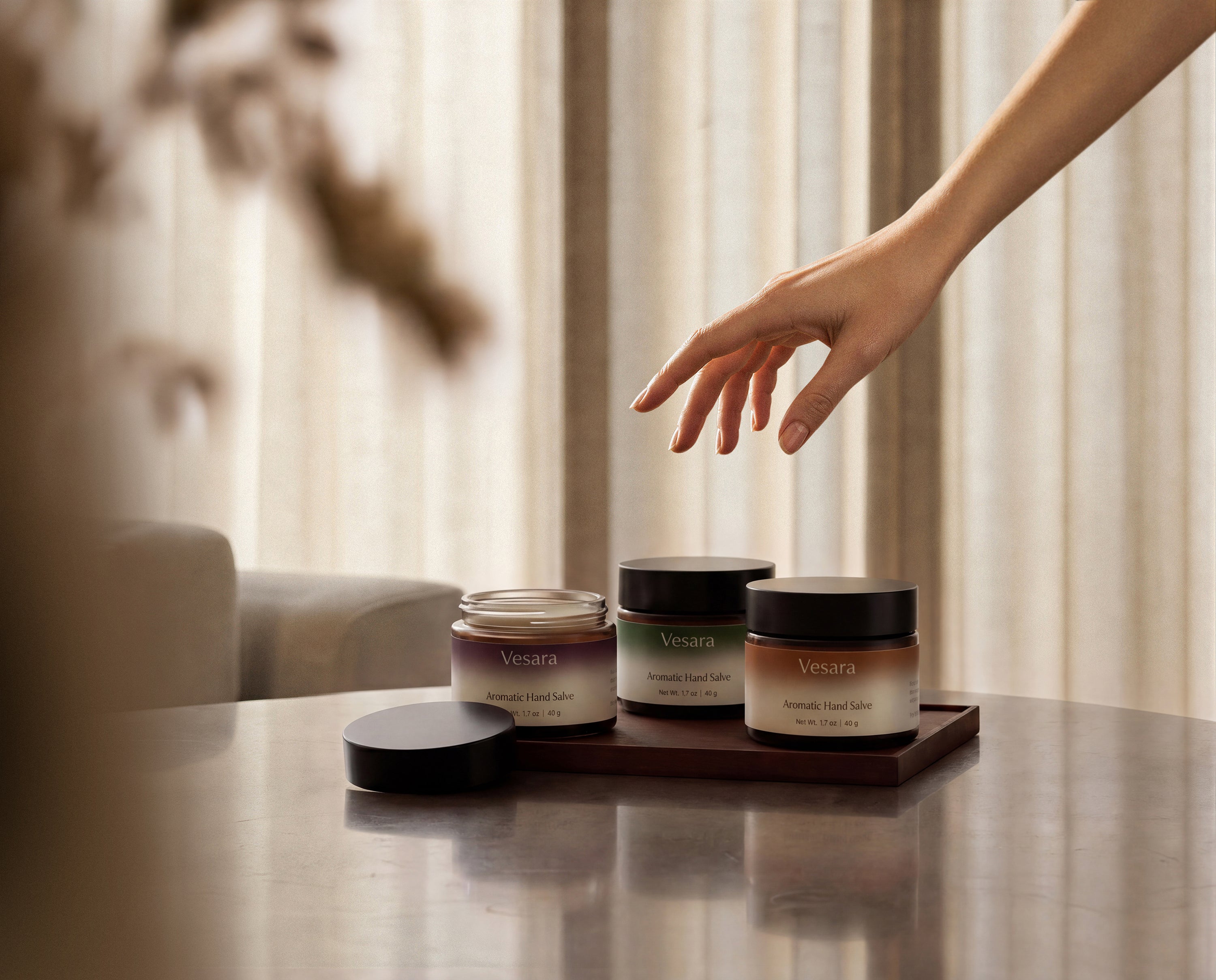 Hand hovering above three Vesara Aromatic Hand Salve jars on warm surface with dramatic shadow lighting