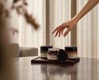 Hand hovering above three Vesara Aromatic Hand Salve jars on warm surface with dramatic shadow lighting