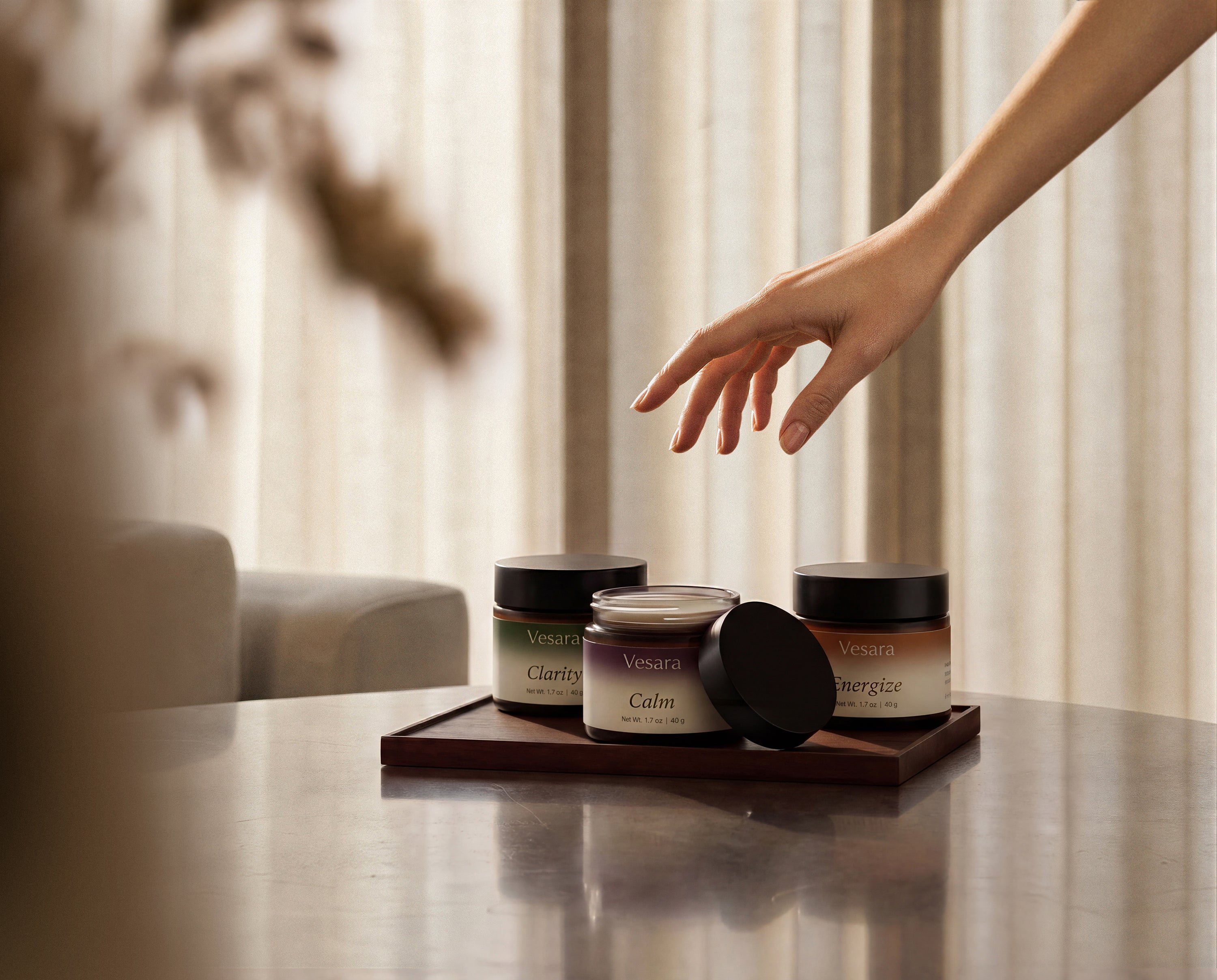 Hand hovering above three Vesara Aromatic Hand Salve jars on warm surface with dramatic shadow lighting
